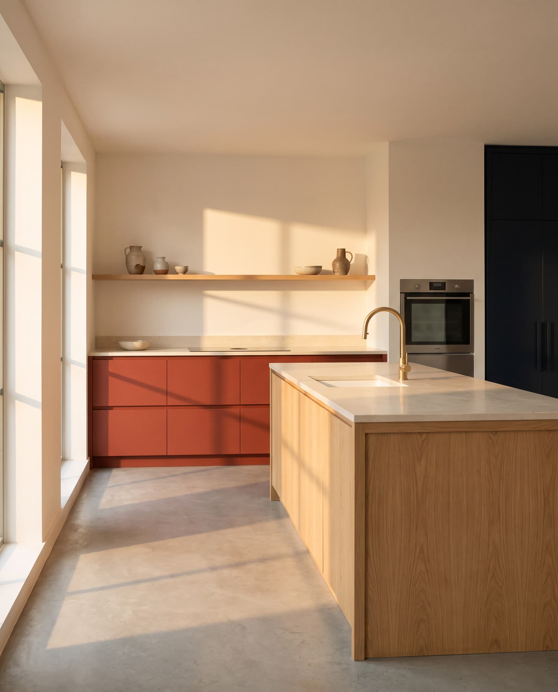 Lege keuken in warm middaglicht met terracottarode onderkasten, een licht eiken kookeiland met stenen blad, een messing kraan en een open eiken wandplank.