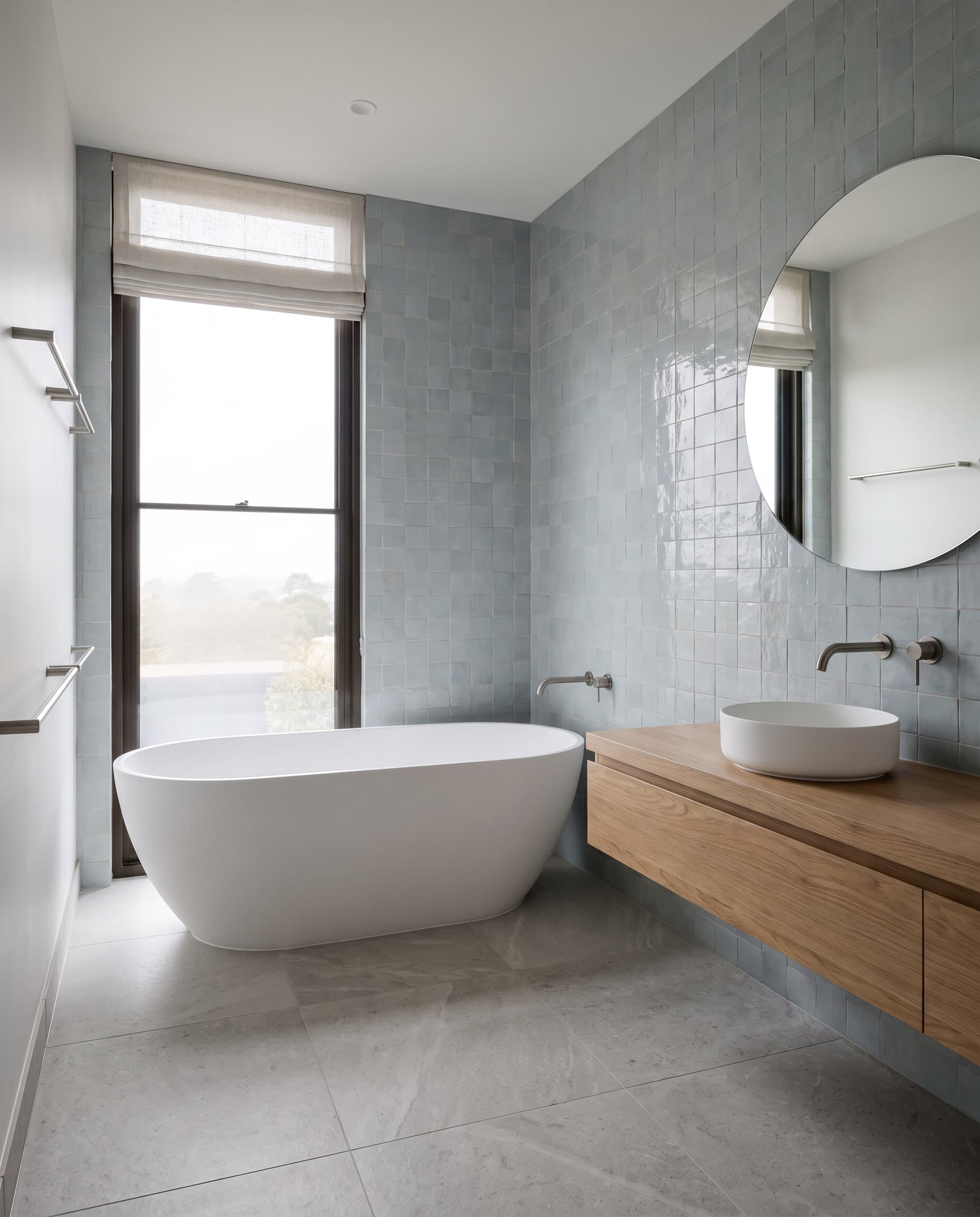 Lege, lichte badkamer met een vrijstaand ovaal bad voor een hoog raam, blauwgrijze handgemaakte wandtegels, een ronde spiegel en een zwevend eiken badmeubel.