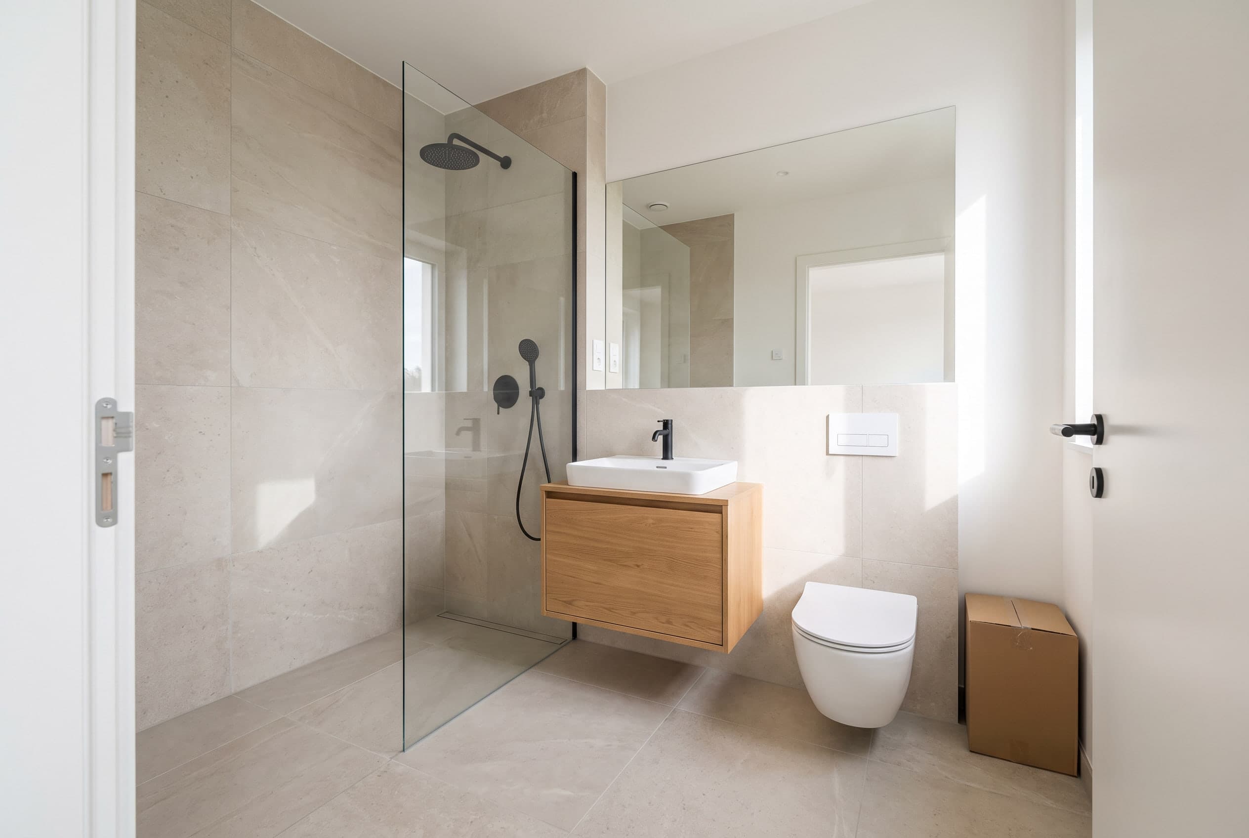 Opleveringsklare badkamer met grote lichte tegels, inloopdouche met glazen wand, zwevend eiken badmeubel en hangtoilet.